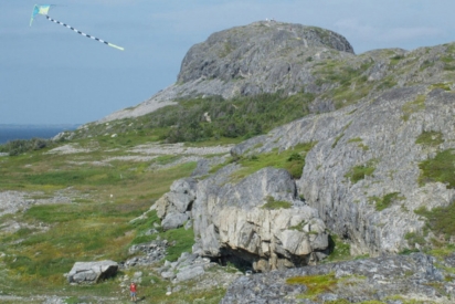 A blue-and-white kite flies in the sky above a rocky, moss-covered hillside landscape, with a distant view of the ocean visible on the left; a small figure in red is faintly visible on the ground near the base of a large boulder in the foreground.
